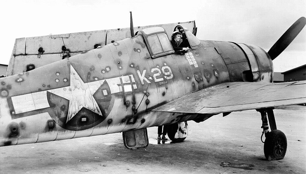 Black-and-white photograph of a World War II fighter plane after combat, its fuselage and wing riddled with bullet holes.